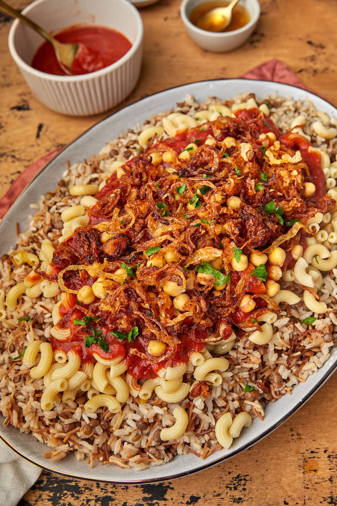 Close up photo of koshari on wooden background with tomato sauce in a container in the background.