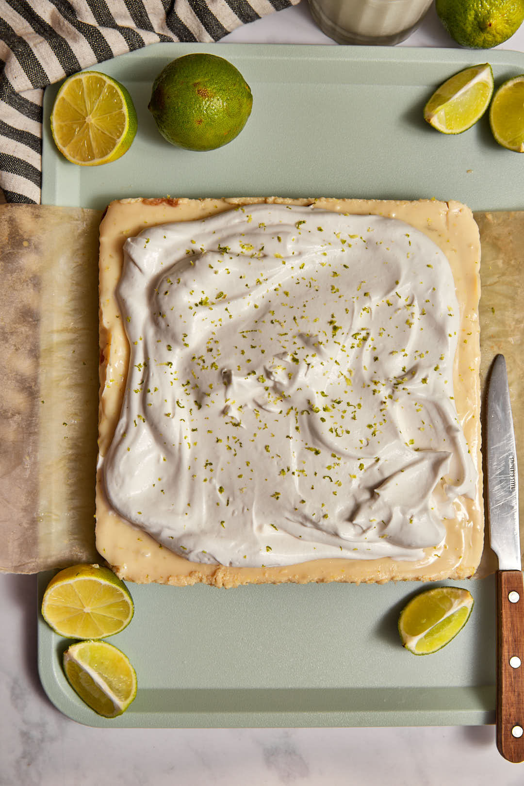 whipped cream spread on key lime pie bars on a mint green tray with a knife on the side.