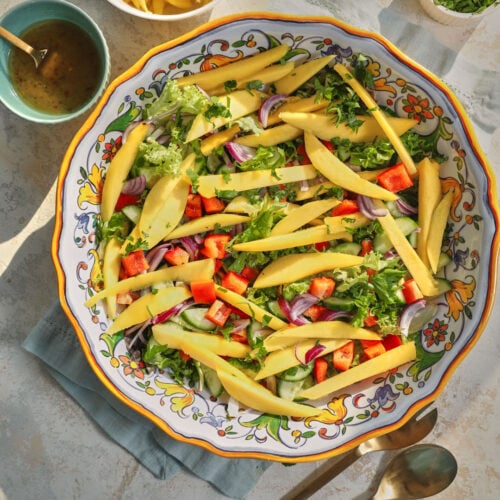 photo of a lettuce and mango salad in a large platter on a blue kitchen linen and on a white background.
