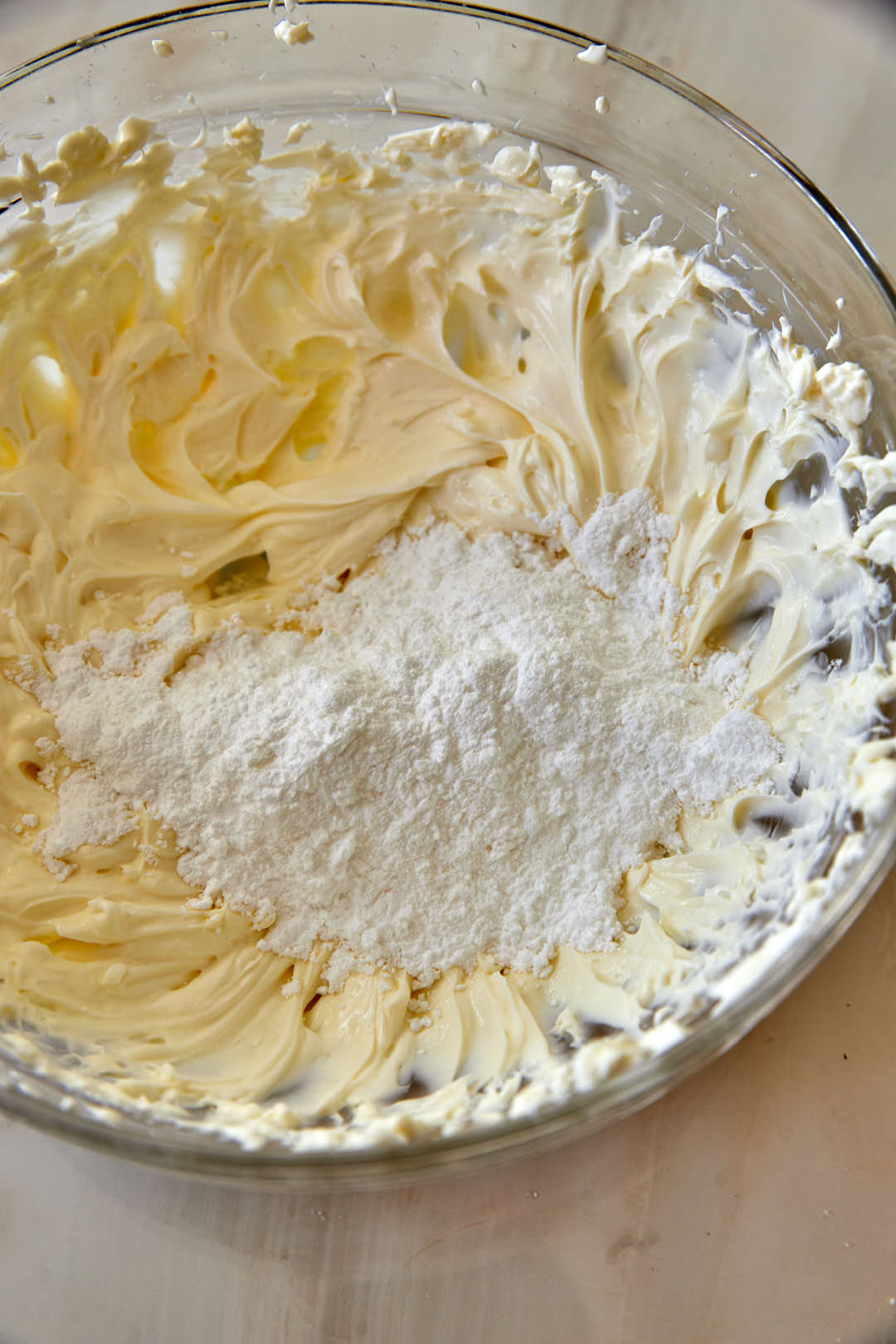 powdered sugar being added to whipped cream cheese in a glass bowl.
