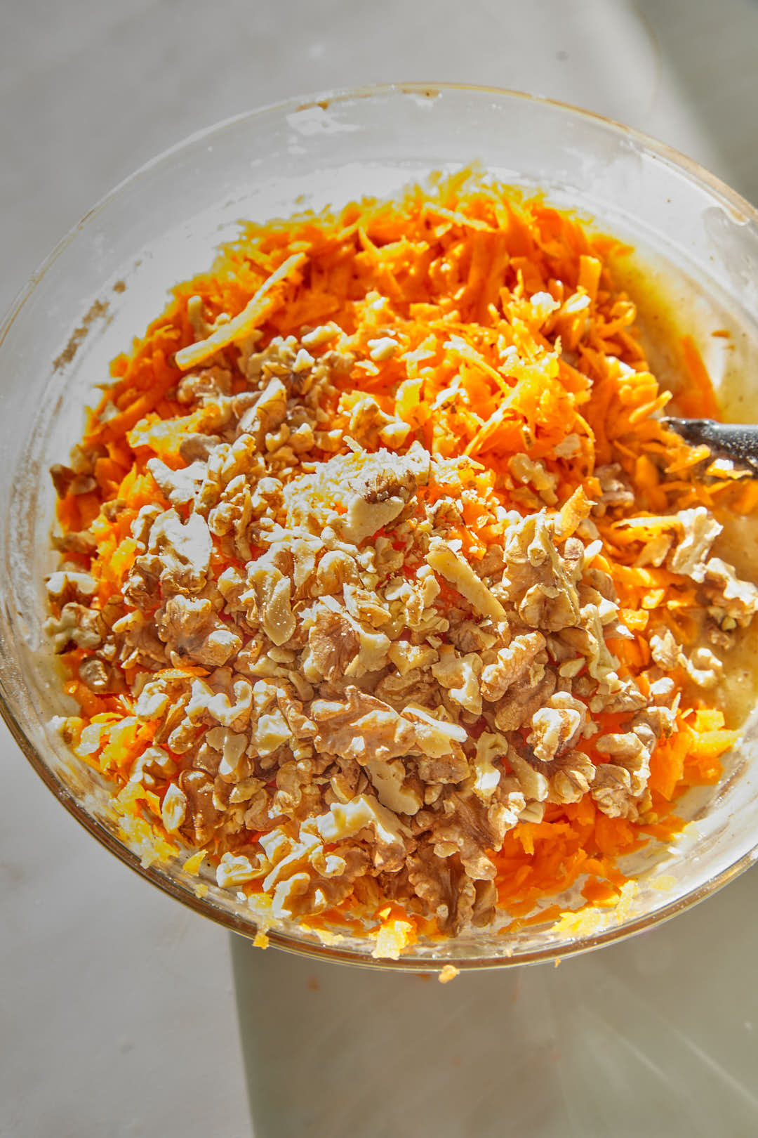 grated carrots and walnuts added to cake batter in a glass bowl.