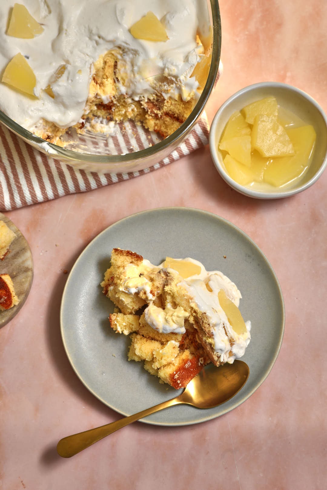 small plate of pineapple trifle with the large trifle in the background, and some pineapple cubes in a small bowl.