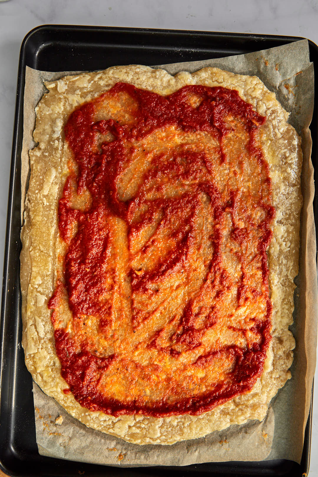 pizza sauce spread onto a rectangle puff pastry pizza dough on a parchment paper lined baking sheet.