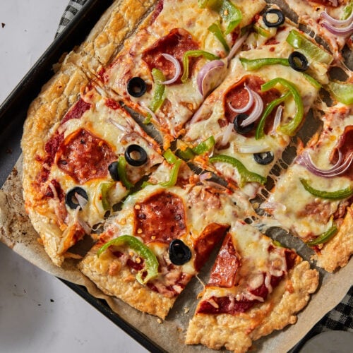 puff pastry pizza on a parchment paper lined baking sheet with pepperoni, olives, and green bell pepeprs, cut into squares.