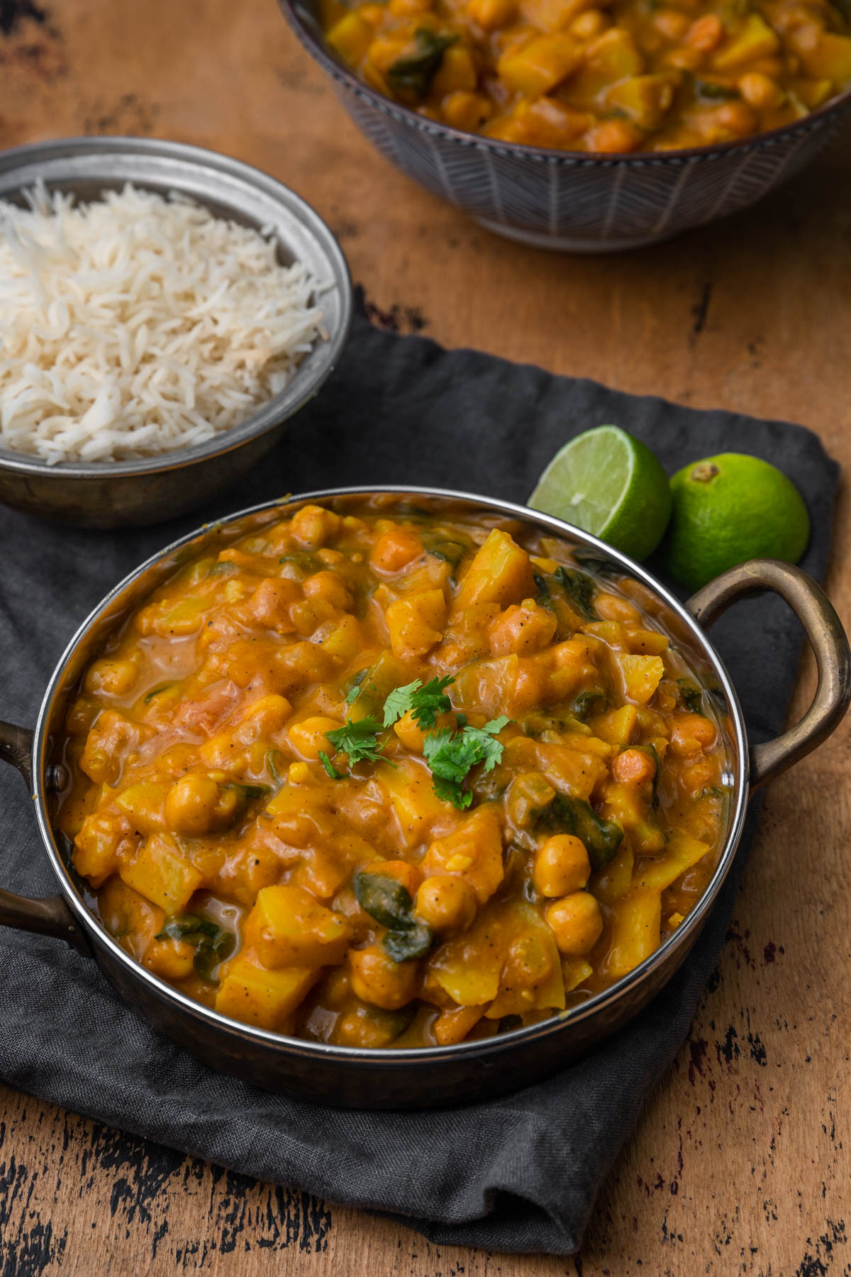 Pumpkin chickpea and sweet potato curry in bronze pan with limes in the background.