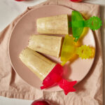three apple popsicles on a pink plate with more apples in the background.