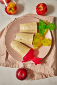 three apple popsicles on a pink plate with more apples in the background.