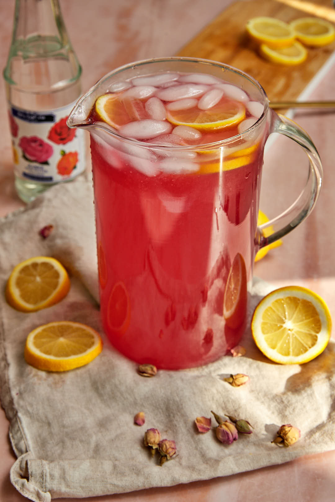 rose lemonade in a pitcher with lemon slices and rose water syrup in the background.