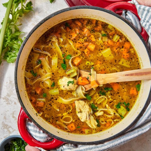 rotisserie chicken noodle soup in a red pot with a wooden spoon and fresh parsley.