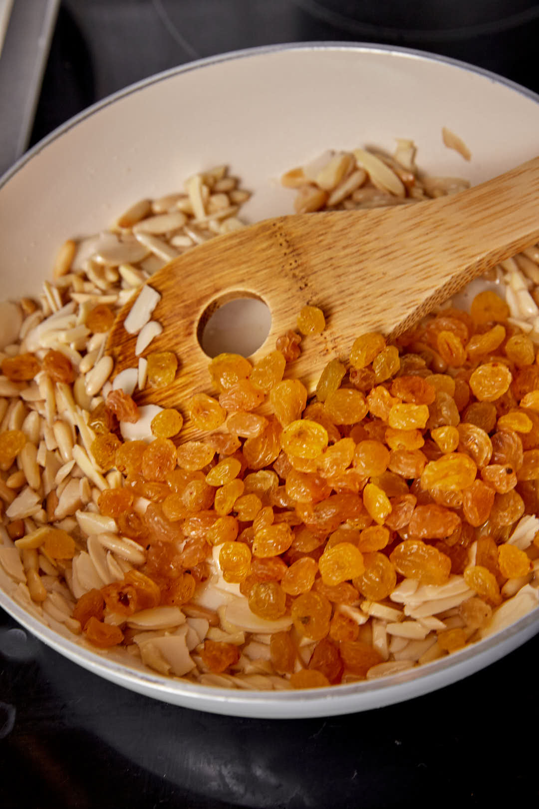 Slivered nuts and golden raisins added to a hot skillet with a wooden spoon.