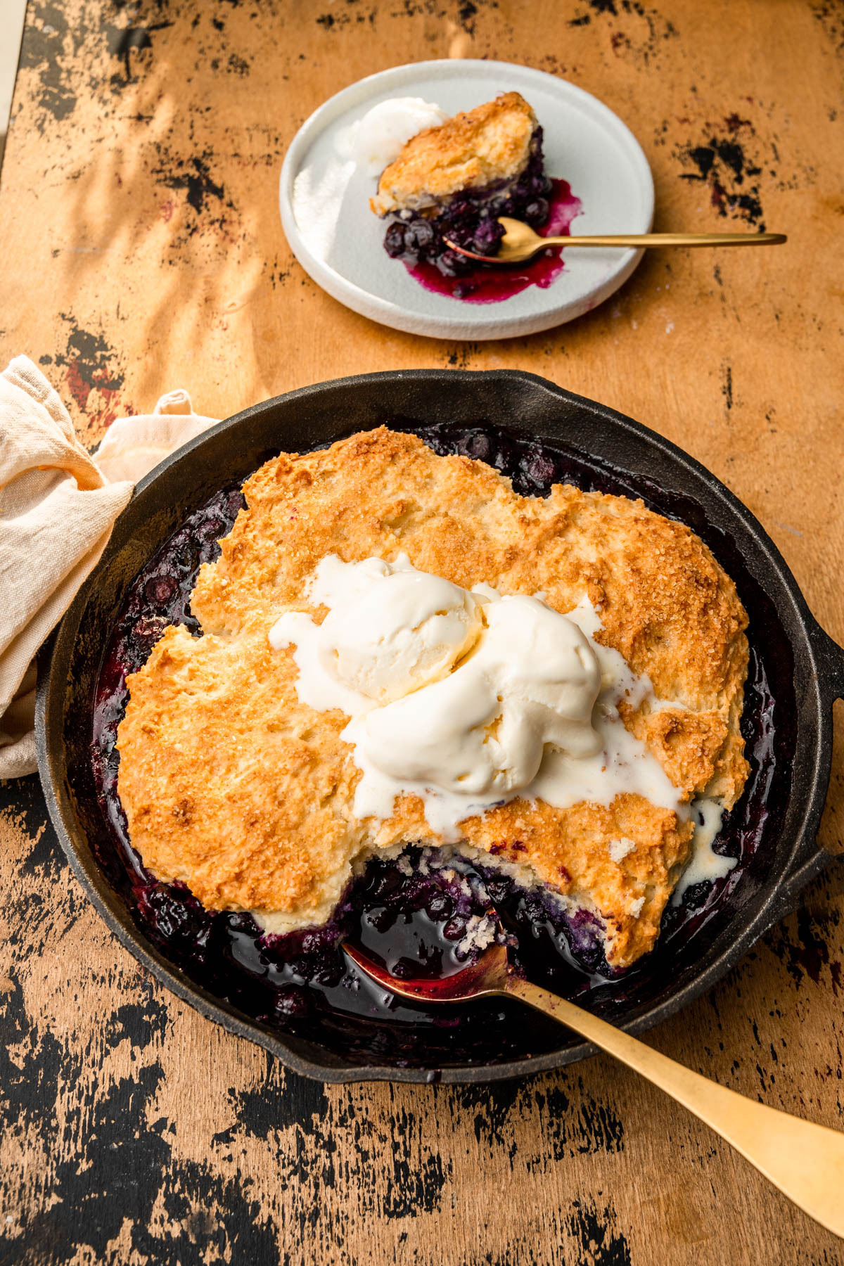 blueberry cobbler in a skillet with ice cream and a golden spoon.