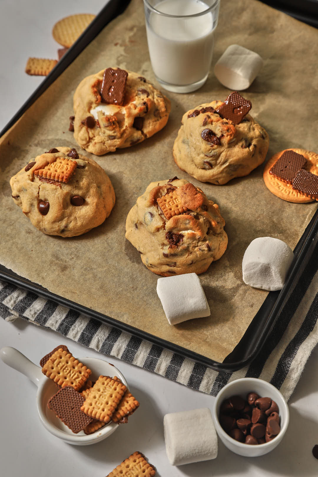 a baking sheet with s'mroed stuffed cookise, marshmallow pieces, and biscuits on a striped kitchen linen.