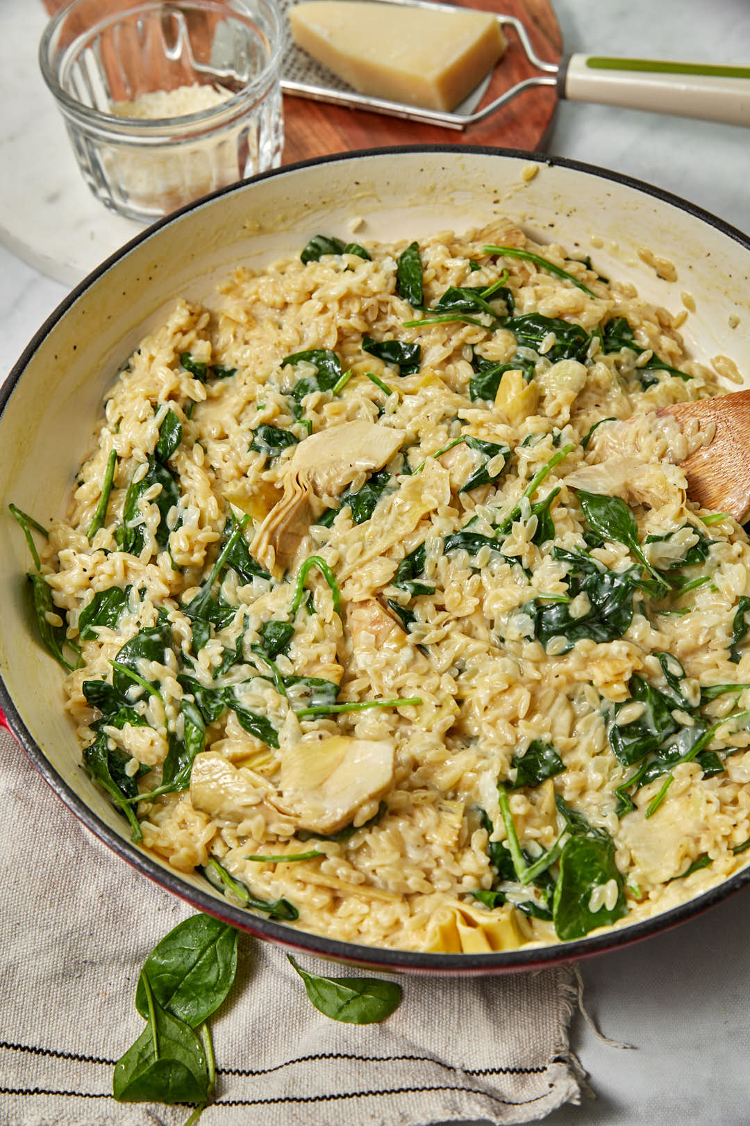 a pot of spinach artichoke orzo with parmesan on a grater in the background and a glass of water in the background.
