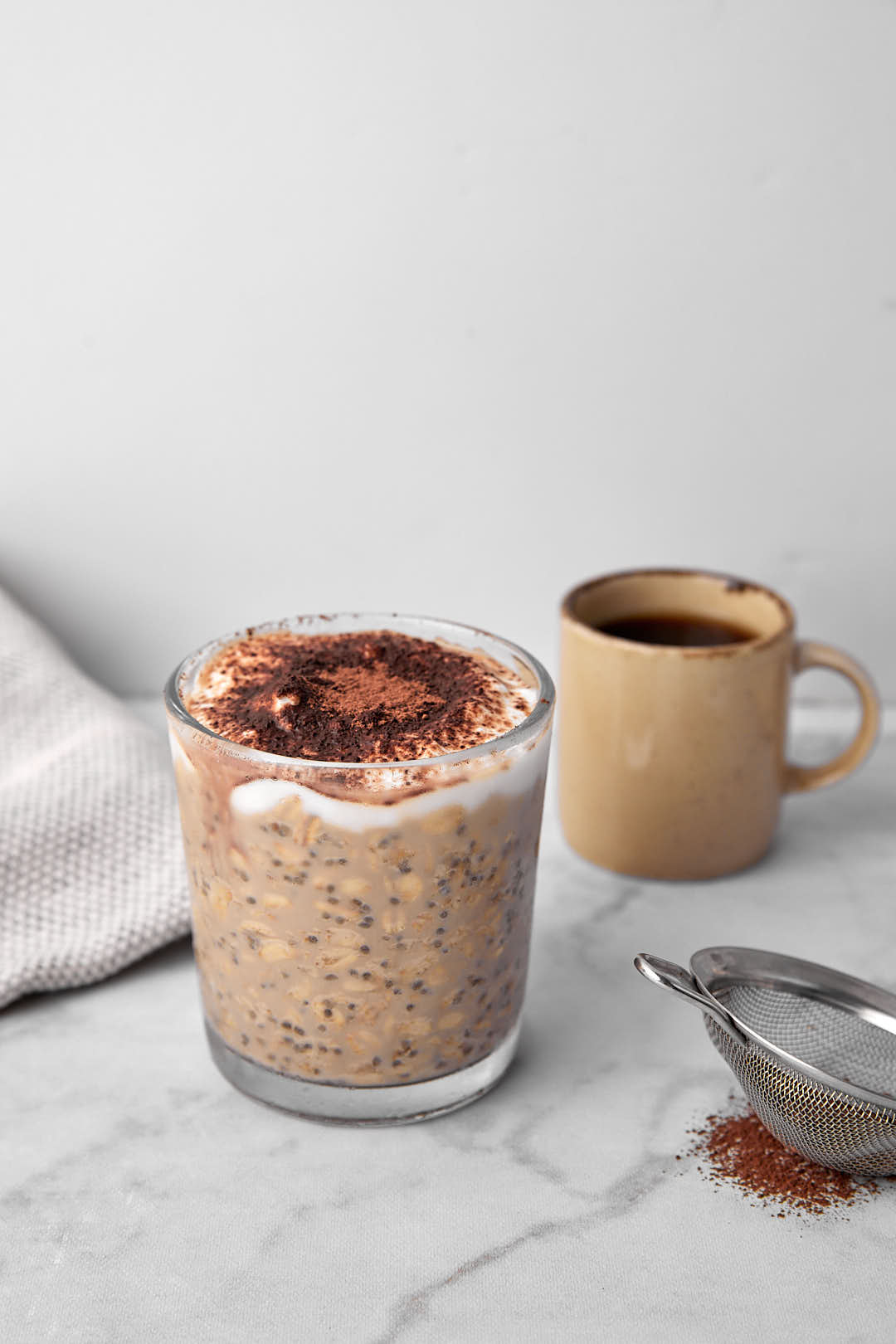 Tiramisu oats in a glass with a cocoa sifter and a mug in the background.