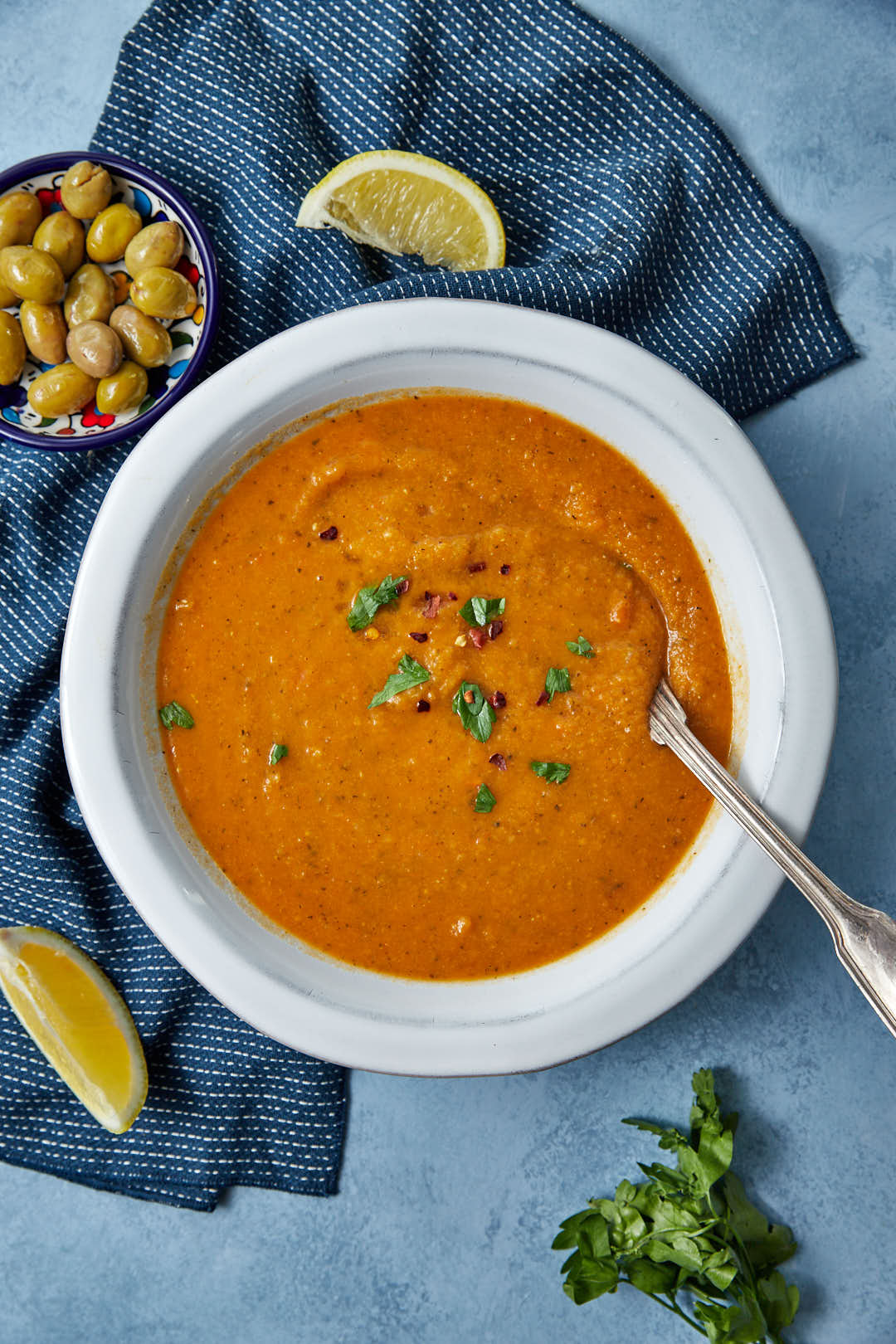 a white bowl of turkish lentil soup, with olives, lemon wedges and parsley on the side on a blue background.