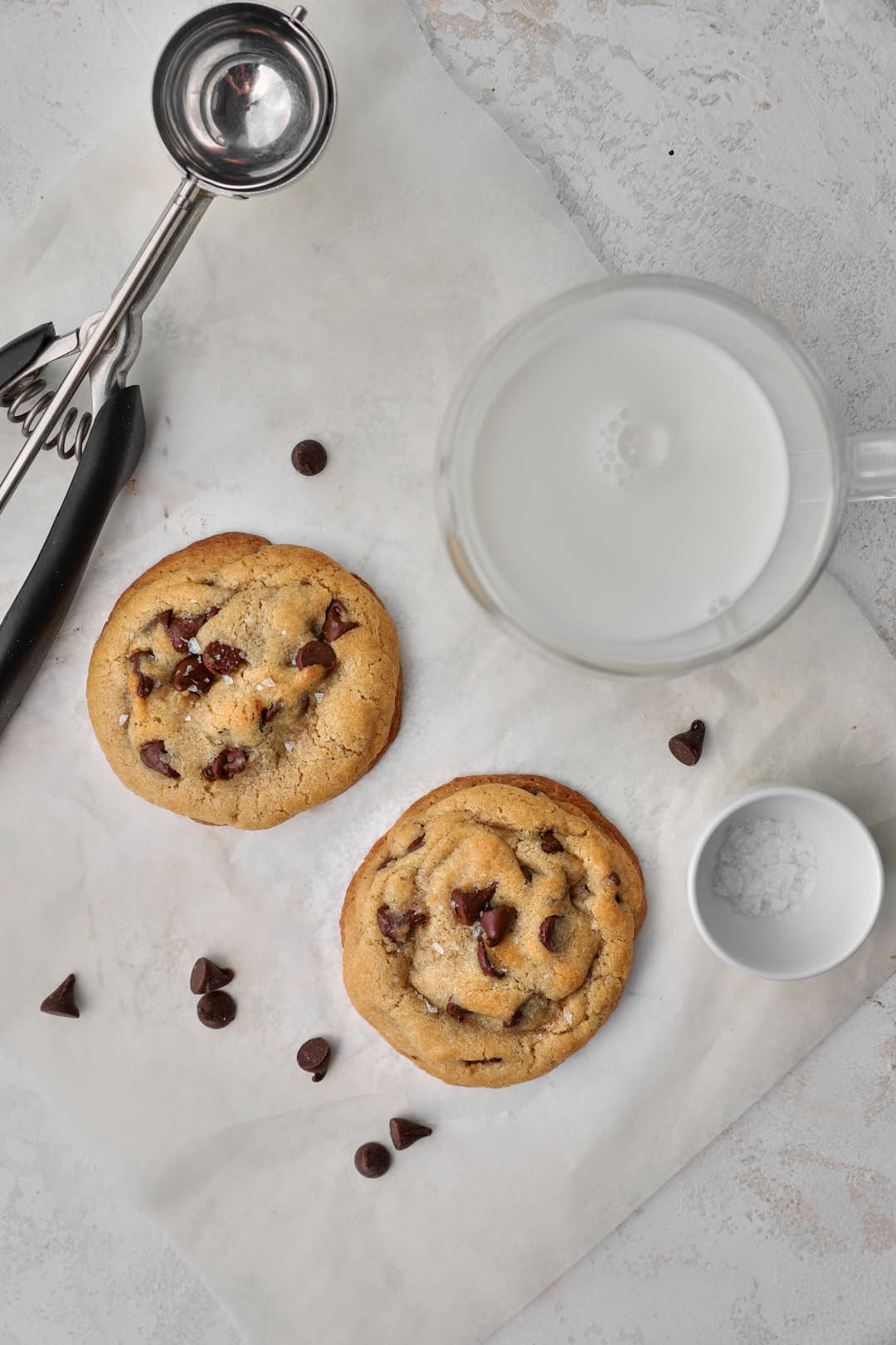 two chocolate chip cookies on parchment paper with a cookie scoop and milk in the background.
