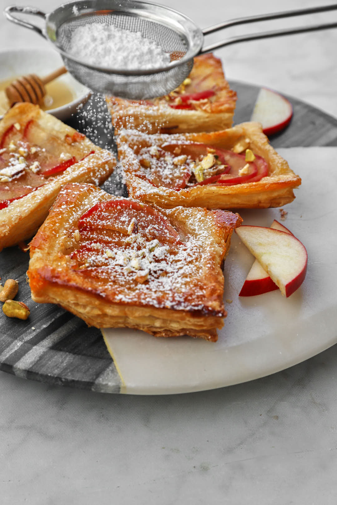 icing sugar being sifted onto apple puff pastry square on a marble cake board.
