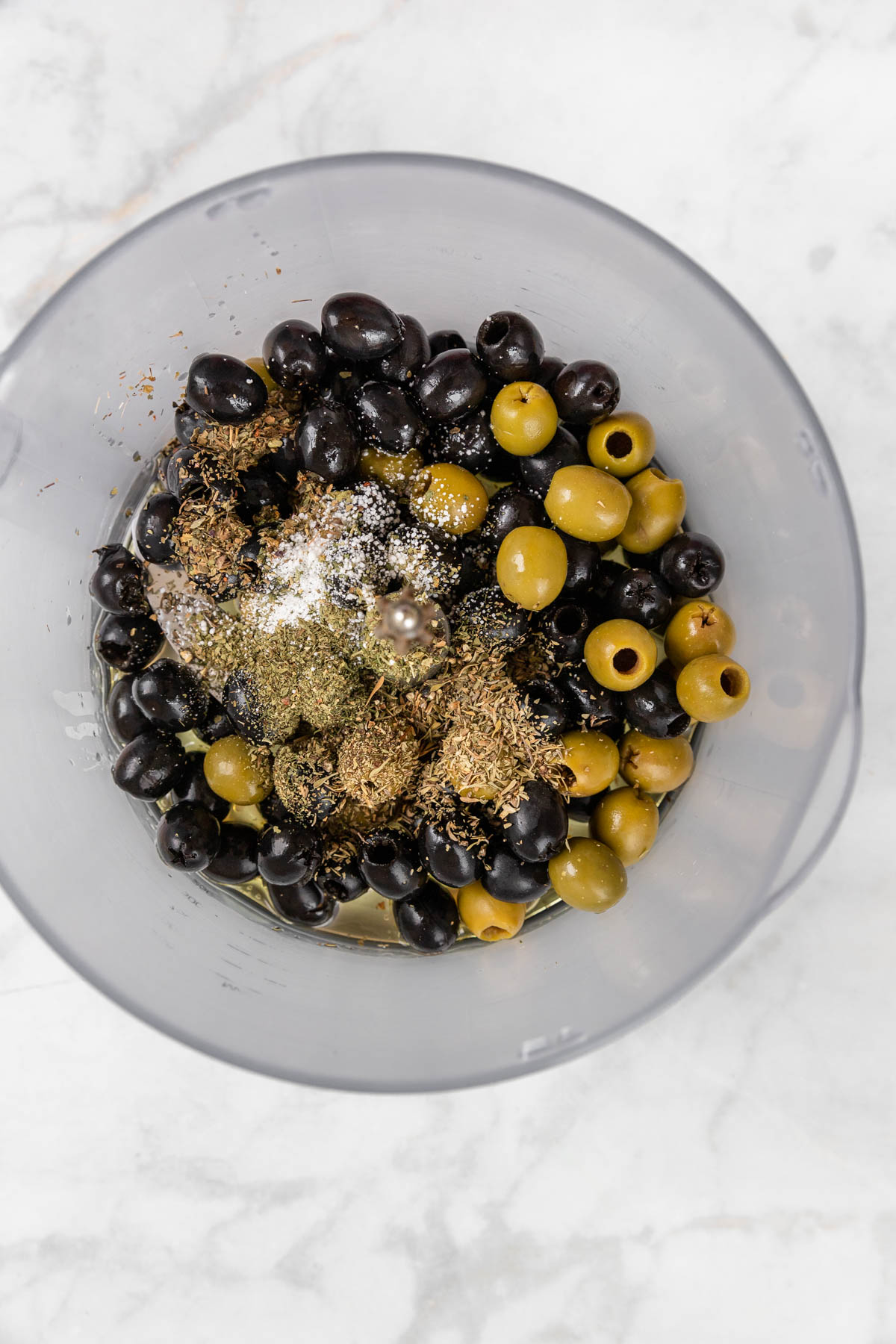 Olives, herbs, olive oil in bowl of a food processor.
