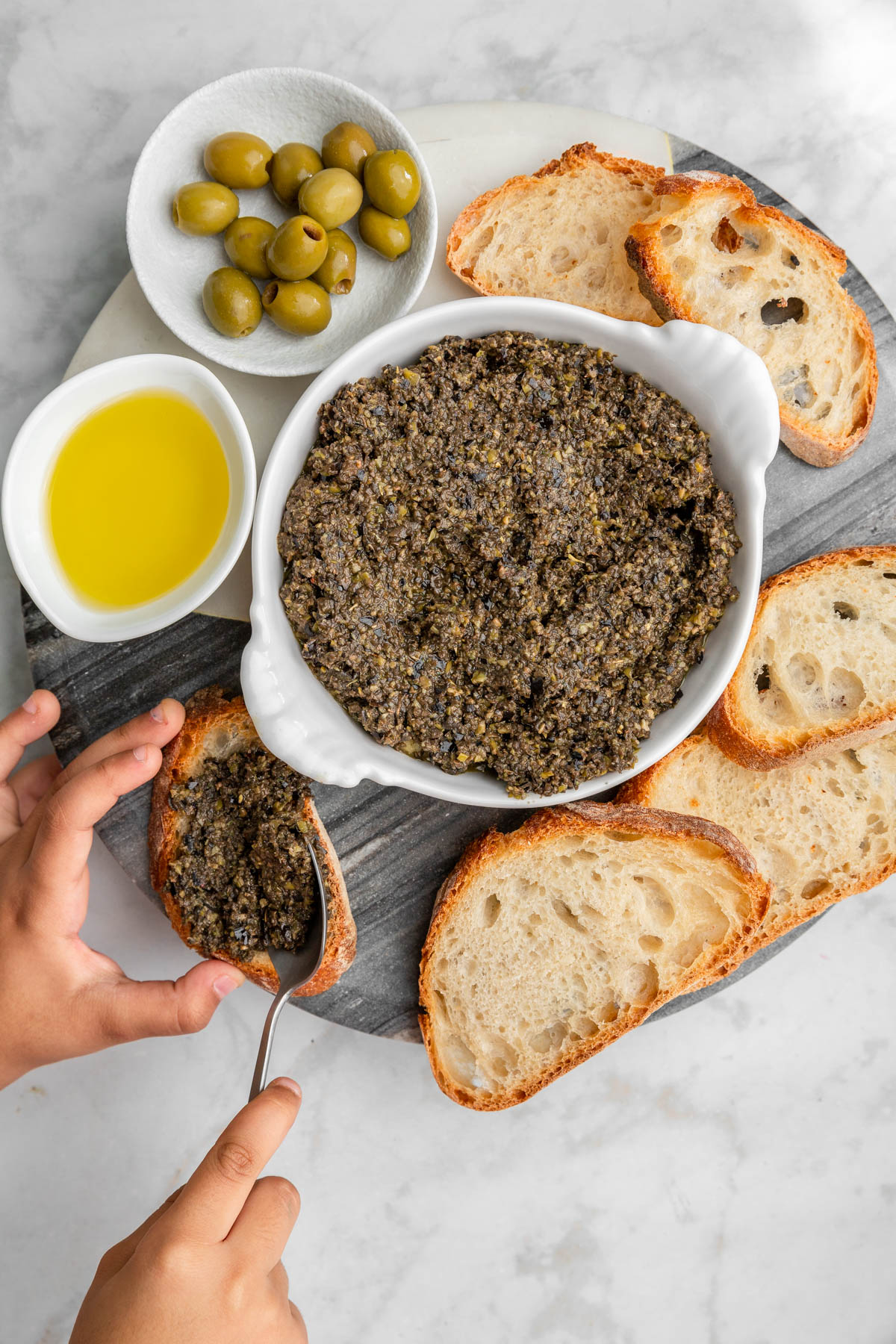 Kids hand holding a piece of bread and spreading olive tapenade on it.