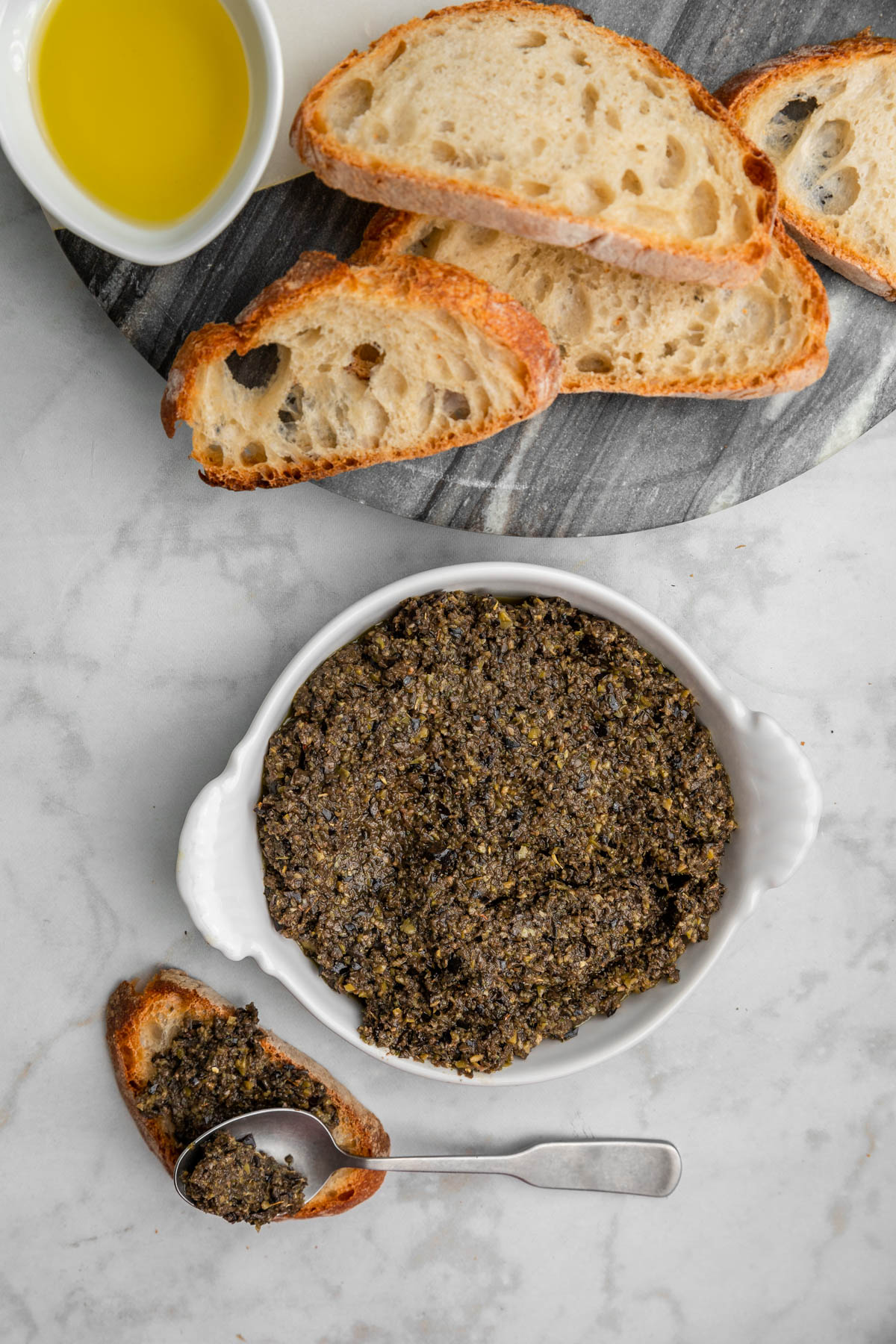 spoon spreading olive tapenade on crusty piece of bread.