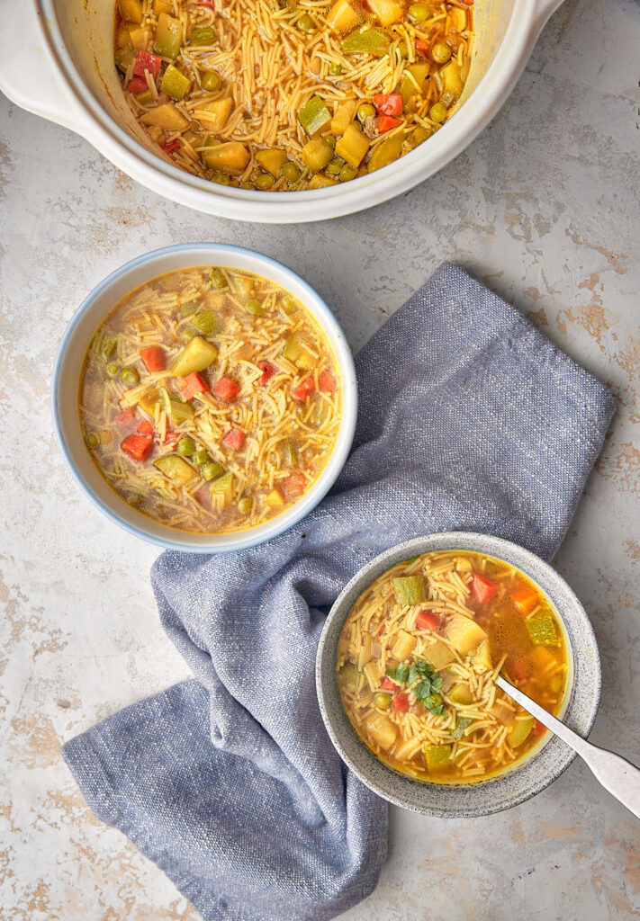 two bowls of vegetable noodle soup on a white background with a blue kitchen towel.