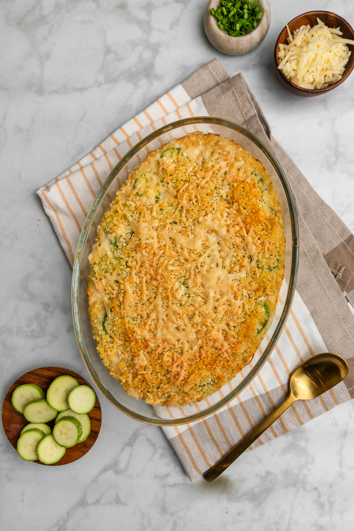 Baked zucchini in a casserole dish with raw zucchinis on the side.